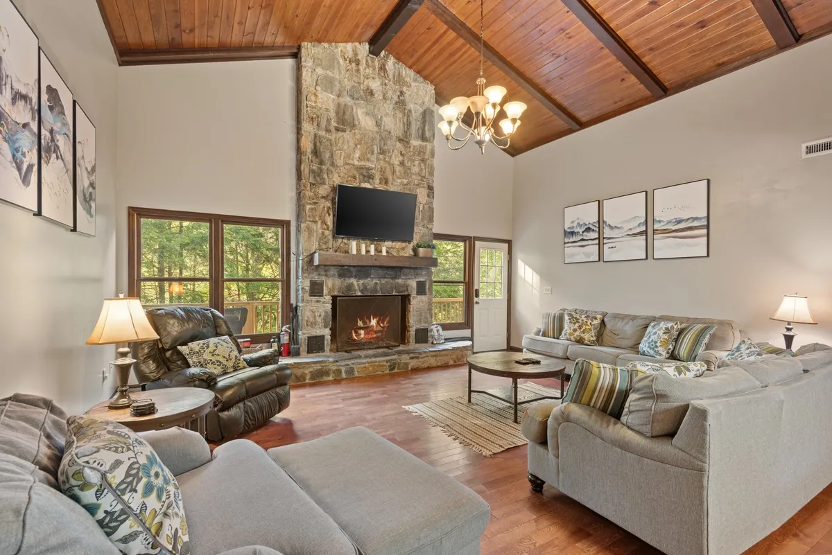 A spacious living room with a striking stone fireplace as its centerpiece, accompanied by large windows that let in natural light. Comfortable seating, including sofas and armchairs, are arranged around the fireplace, creating a warm and welcoming environment. Overhead, a rustic chandelier and wood accents add a mountain-lodge character to the space.