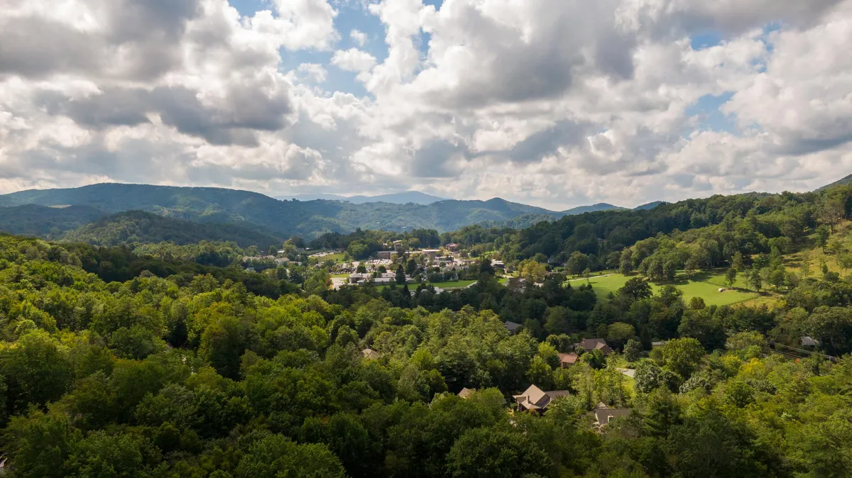 A sweeping view captures rolling hills and lush greenery dotted with wildflowers, with the quaint town of Banner Elk nestled in the valley below. The scene is tranquil, with a cloudy sky hinting at the area's changing seasons and the natural beauty that surrounds the town.