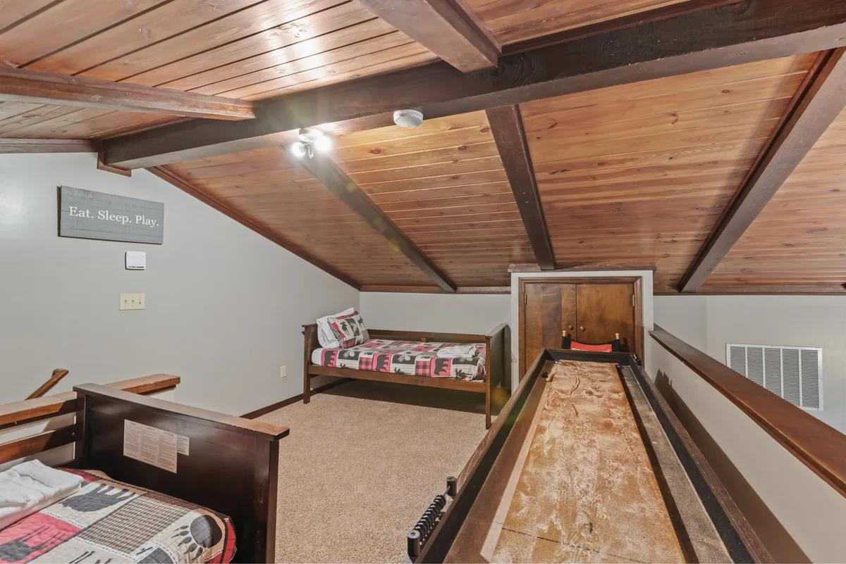 This inviting loft space features wooden beams, a playful slogan sign, daybeds for lounging, and a shuffleboard table taking center stage. The overall atmosphere is relaxed and fun, perfect for friendly games or chilling out with friends.