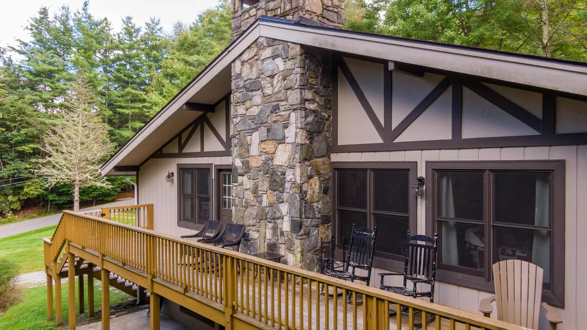 The image showcases the inviting exterior of a mountain lodge, featuring a sturdy stone chimney, a spacious wooden deck lined with classic rocking chairs, and surrounded by lush natural scenery. The setting is perfect for relaxing, enjoying mountain air, and watching sunsets over the pines.