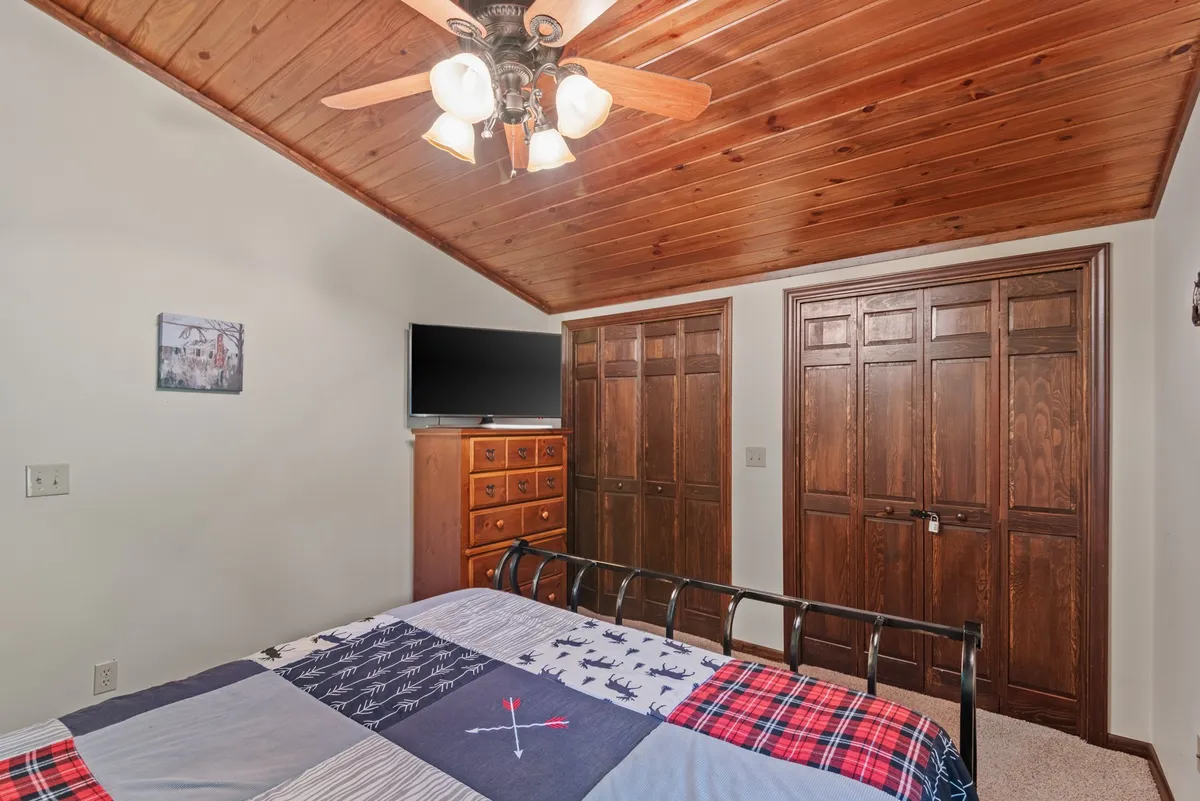 A king bedroom featuring a bed with a patterned comforter, warm wooden ceiling, TV, and wooden closet doors. The room is neatly arranged for both comfort and privacy, with soft lighting and cozy decor.