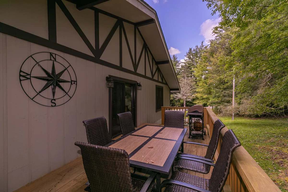The photo features a large wooden dining table on an inviting outdoor patio, surrounded by greenery. Comfortable outdoor seating encourages relaxation, while decorative touches like a compass wall accent evoke a sense of exploration and adventure. The table is perfect for enjoying coffee in the morning or gathering in the evening.