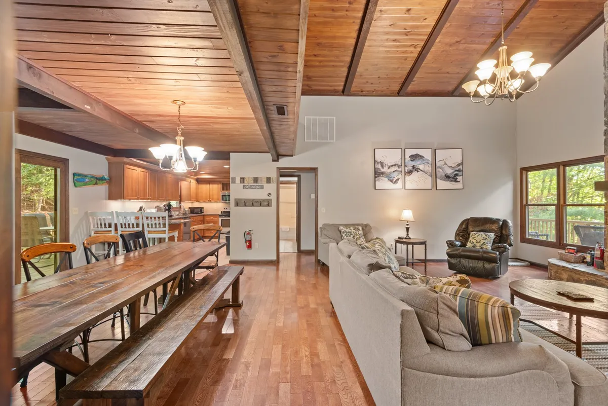 An open-plan great room featuring a large wooden dining table set for a group, with a comfortable seating area nearby. The kitchen area is visible in the background, showcasing ample space for simultaneous cooking and socializing. Decorative artwork and warm lighting enhance the welcoming mood.