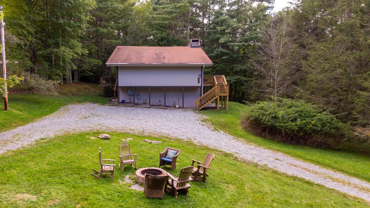 A view of the home's exterior featuring a spacious fire pit area surrounded by comfortable seating, the main house in the background, and a pathway leading toward the gathering spot. The setting is surrounded by natural greenery, perfect for group gatherings and outdoor enjoyment.
