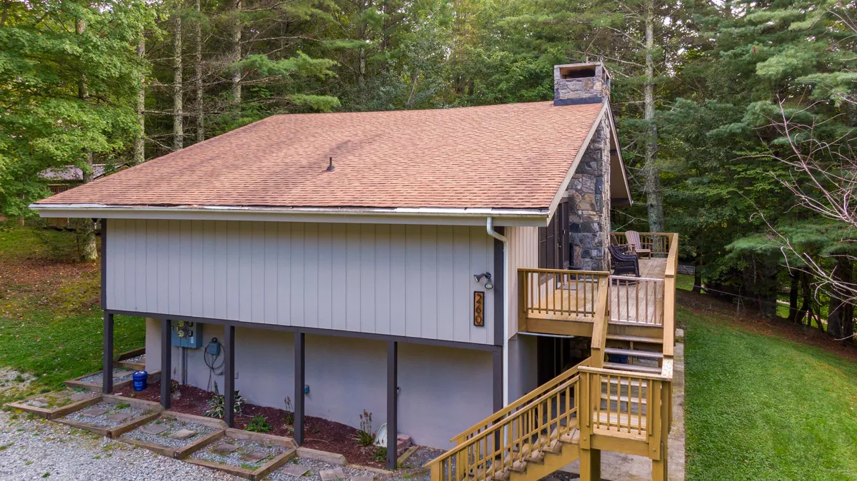 A cozy exterior view of a wooden deck with a stone chimney, set among surrounding trees. The space is perfect for outdoor dining, barbecuing, or simply enjoying the natural surroundings at any time of day.